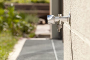 A frost free spigot protrudes from the wall of a house in the backyard. In the background, you can see greenery and a porch.