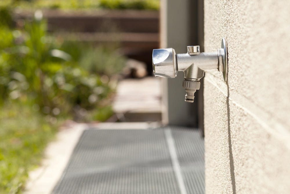 A frost free spigot protrudes from the wall of a house in the backyard. In the background, you can see greenery and a porch.
